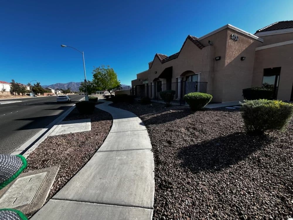 Sidewalk view of a commercial building with shrubs and mountains under a clear blue sky.