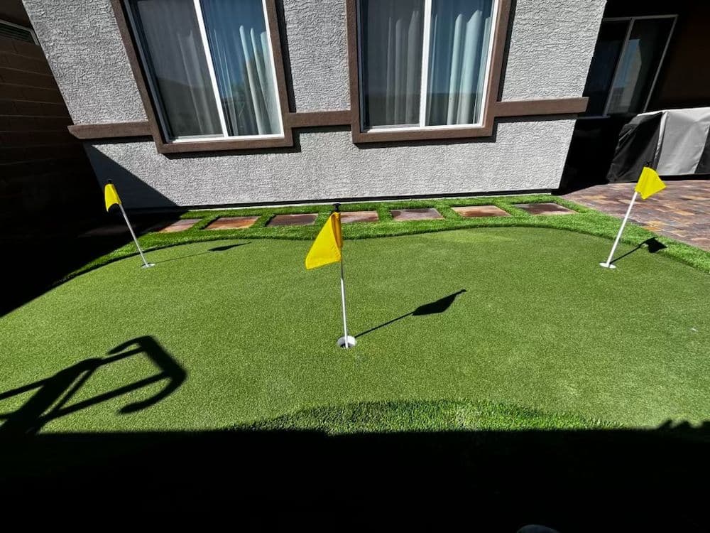 Backyard putting green with yellow flags and artificial turf, bright sunlight illuminating the area.