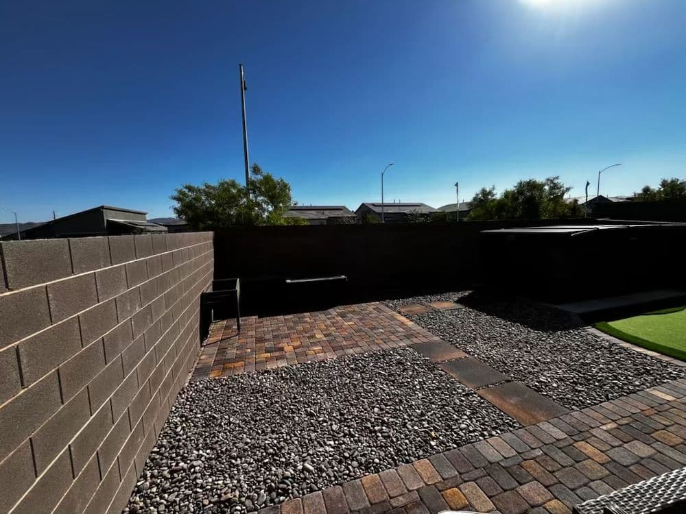 Backyard patio with pavers and gravel, fenced area under clear blue sky.