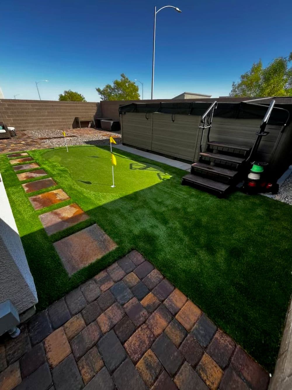 Artificial turf putting green with yellow flags, surrounded by stone pathways and a hot tub.