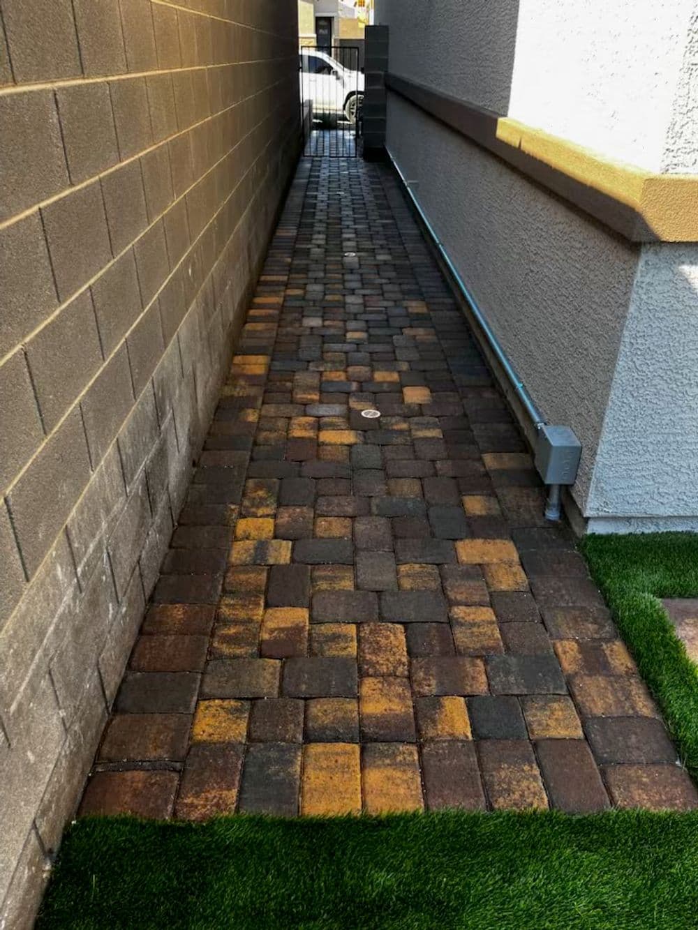 Narrow walkway with brick pavers and artificial grass beside a building exterior.