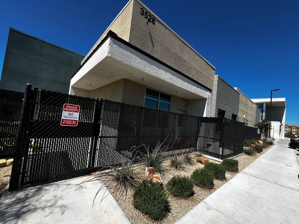 Modern commercial building with security fence and landscaped entrance in a clear blue sky.