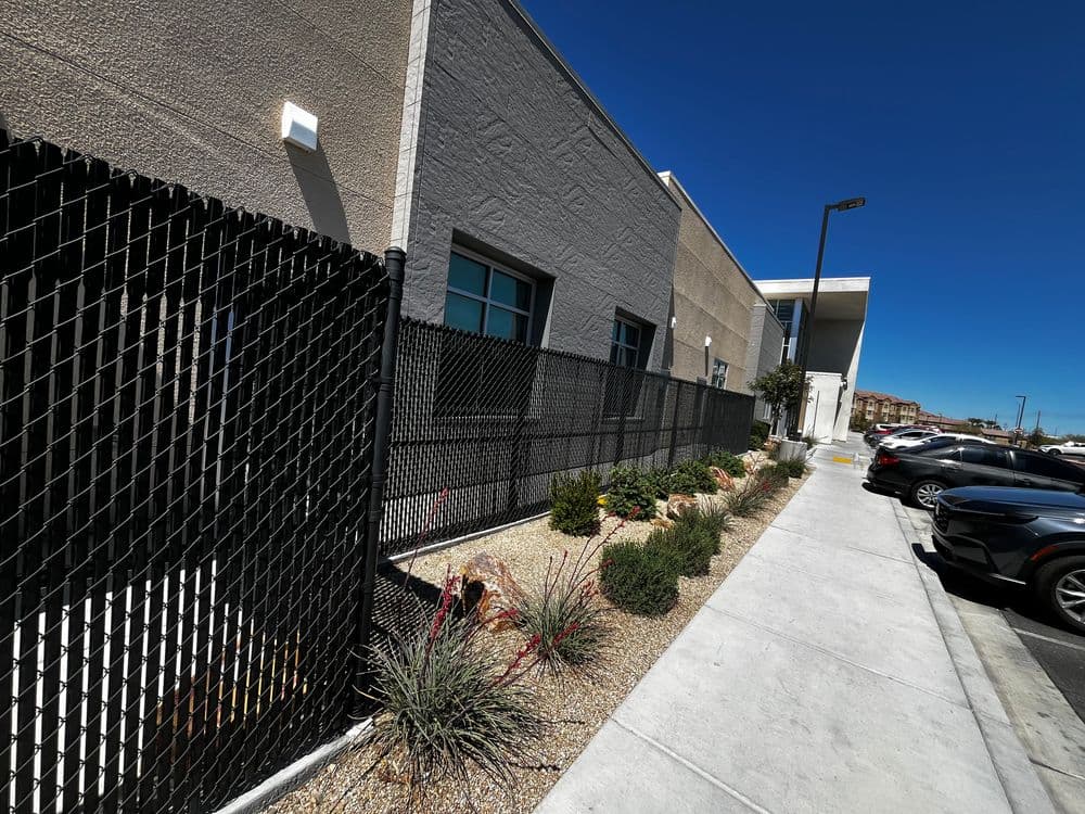 Modern building exterior with black fence and landscaped sidewalk in clear blue sky.